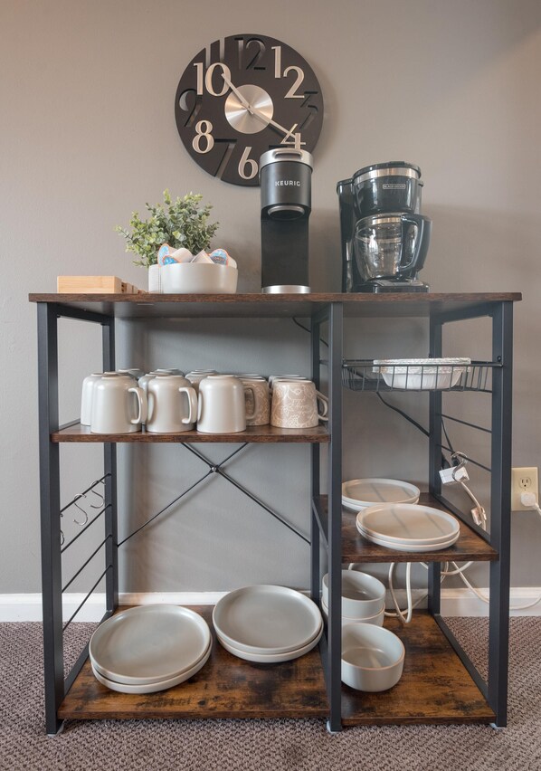 Una zona de cocina con cafetera, tazas y platos sobre una estantería metálica.