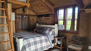 Une chambre dans une cabane en bois, avec un lit, une table de chevet, une chaise, une échelle et une fenêtre avec des rideaux.