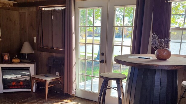 Un intérieur en bois avec une grande barrique, une table ornée d’un vase et une vue sur un lac, visible à travers des portes vitrées.