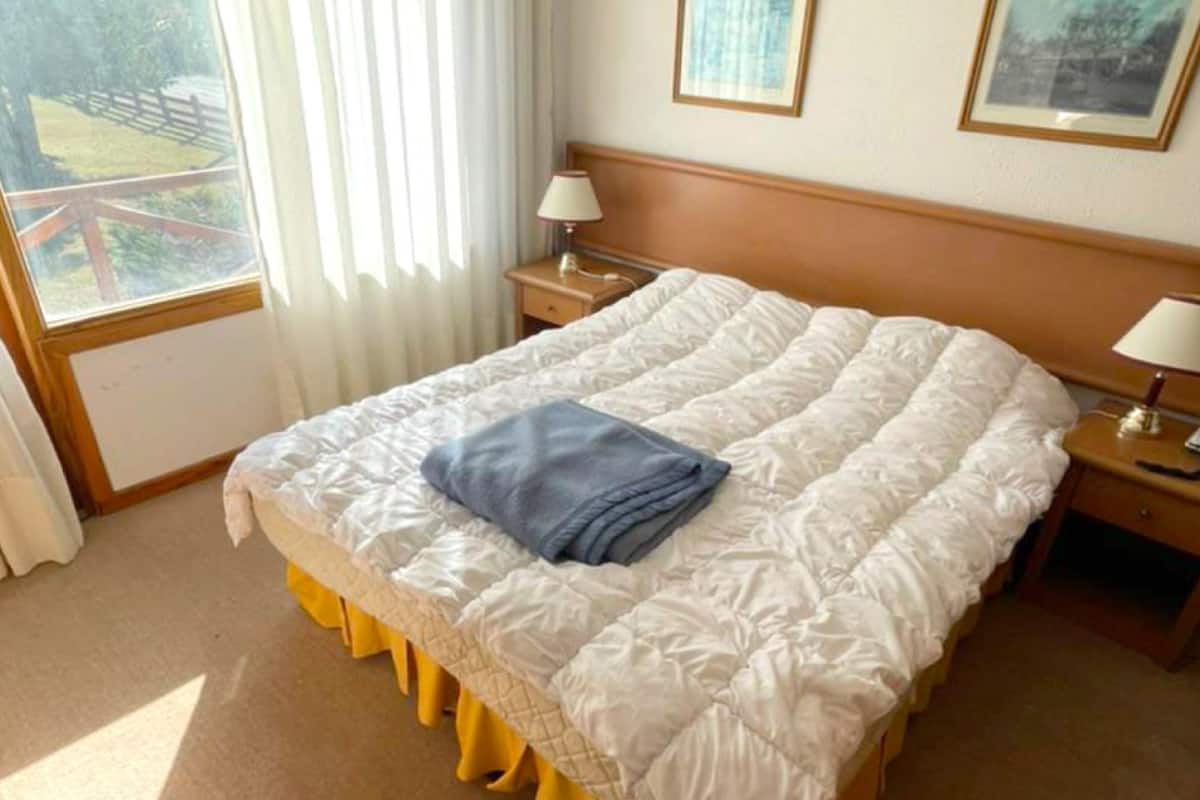 A hotel room with a large bed, two bedside tables with lamps, a window with curtains, and framed pictures on the wall.