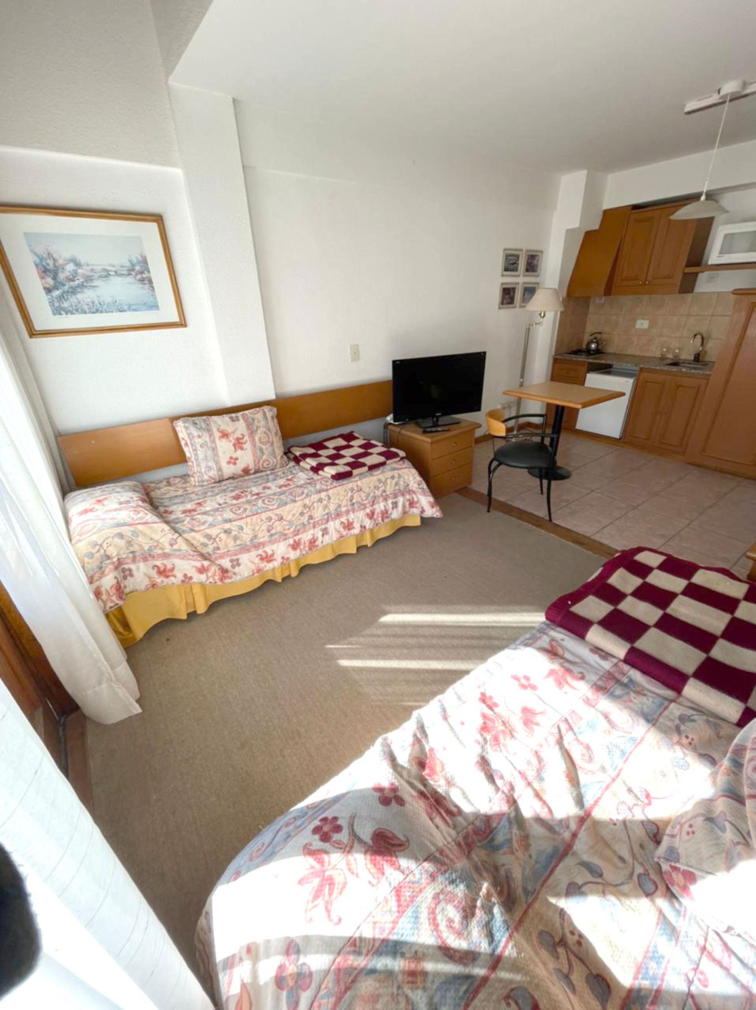 A bedroom with two beds, a TV, a desk, and a kitchenette.