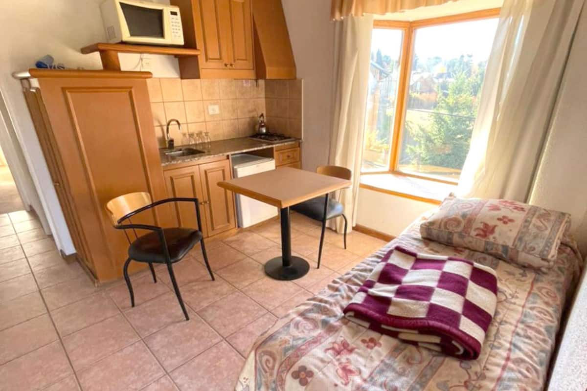 A compact room with a kitchenette, a dining area, and a bed with a patterned blanket.