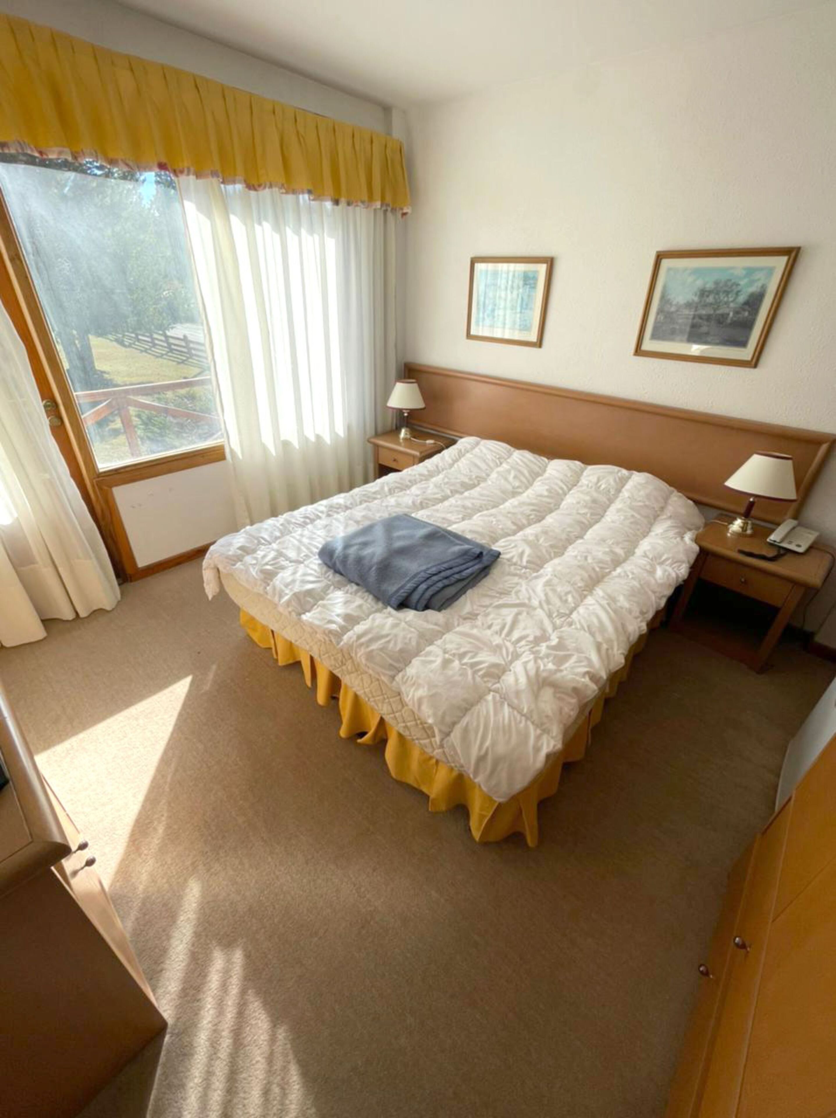 A hotel room with a large bed, two bedside tables with lamps, a window with curtains, and two framed pictures on the wall.