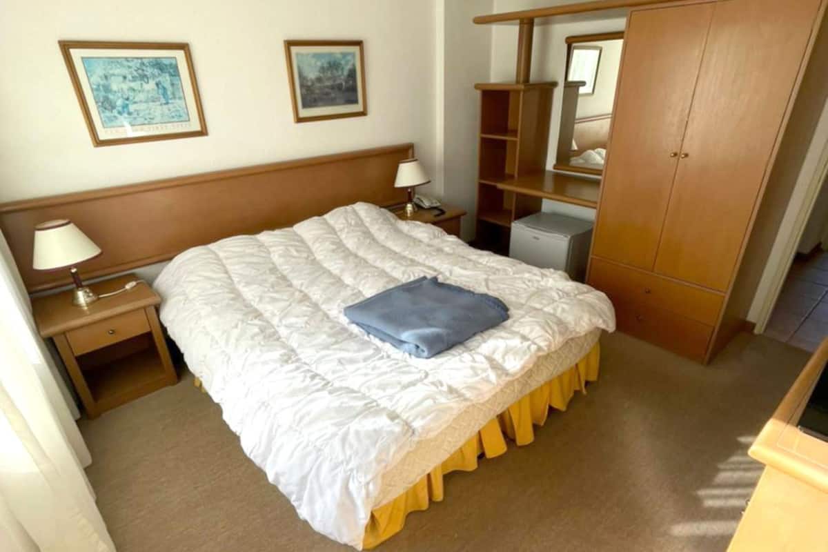 A bedroom with a large bed, a nightstand, a lamp, a wardrobe, and two framed pictures on the wall.