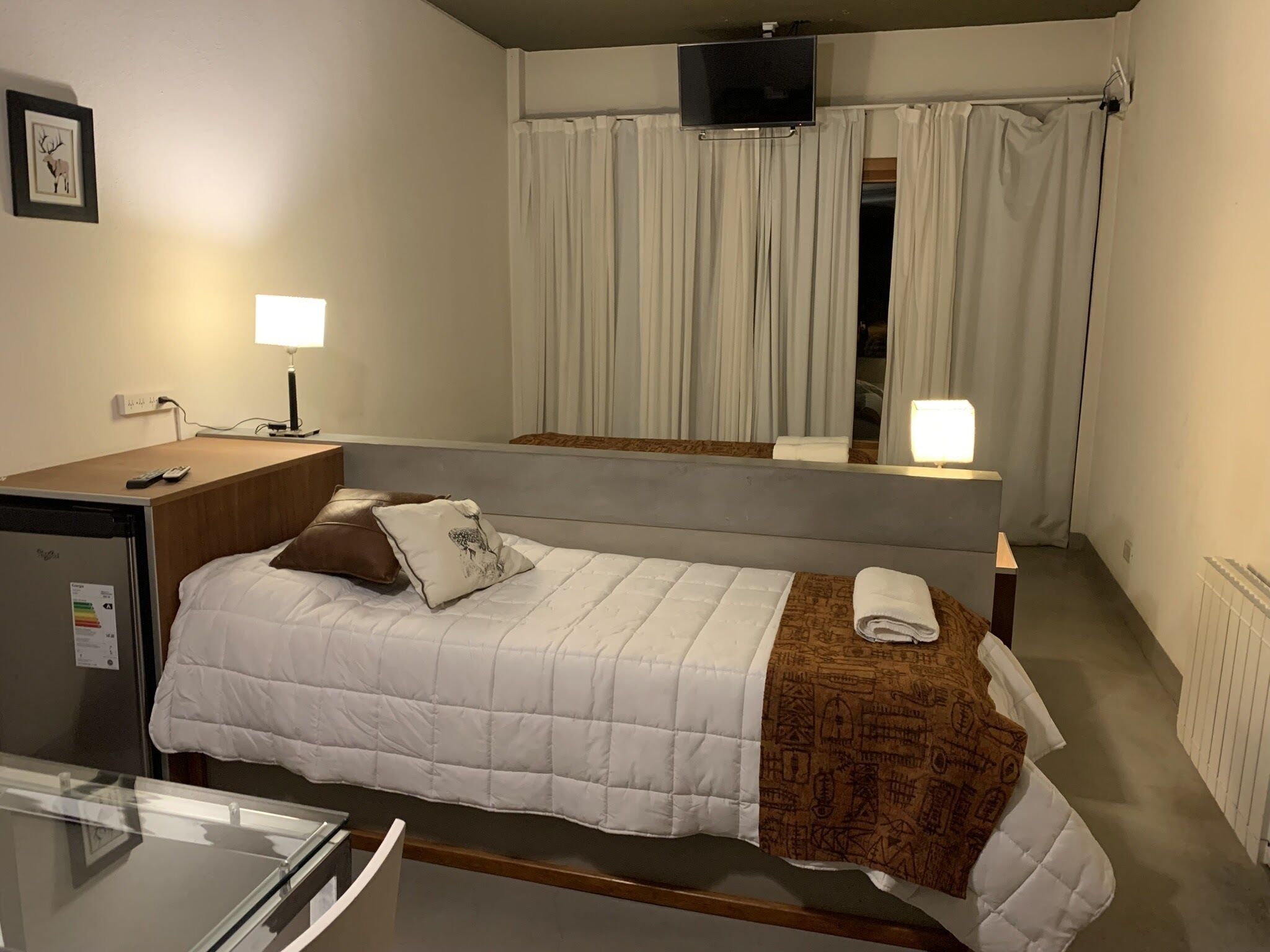 Habitación de hotel con una cama individual, un escritorio con ordenador, un televisor sobre la cama y una ventana con cortinas.