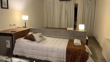 Habitación de hotel con una cama individual, un escritorio con ordenador, un televisor sobre la cama y una ventana con cortinas.