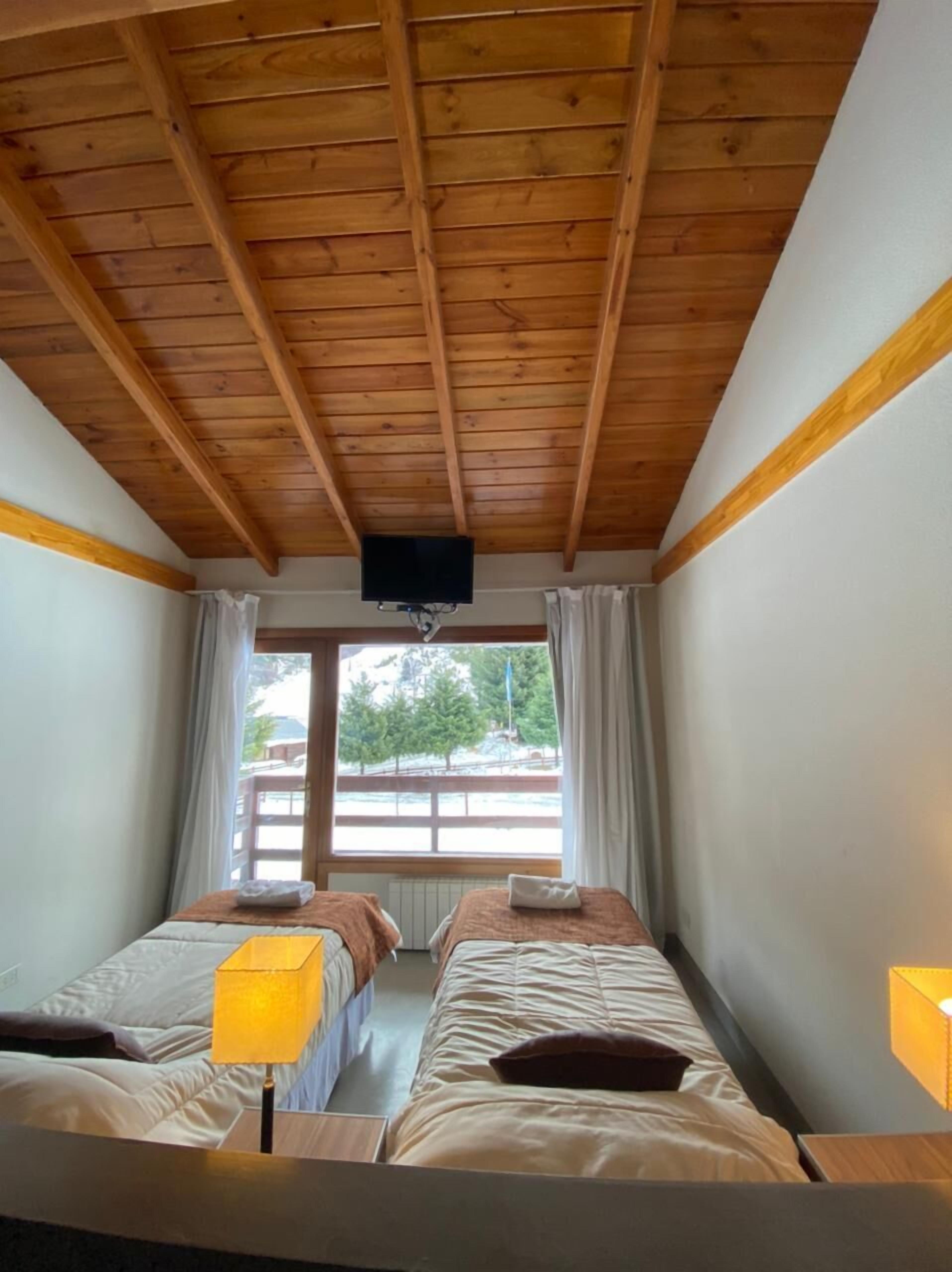 A room with wooden ceiling beams, two beds with pillows, a window with curtains, and a TV mounted on the wall.