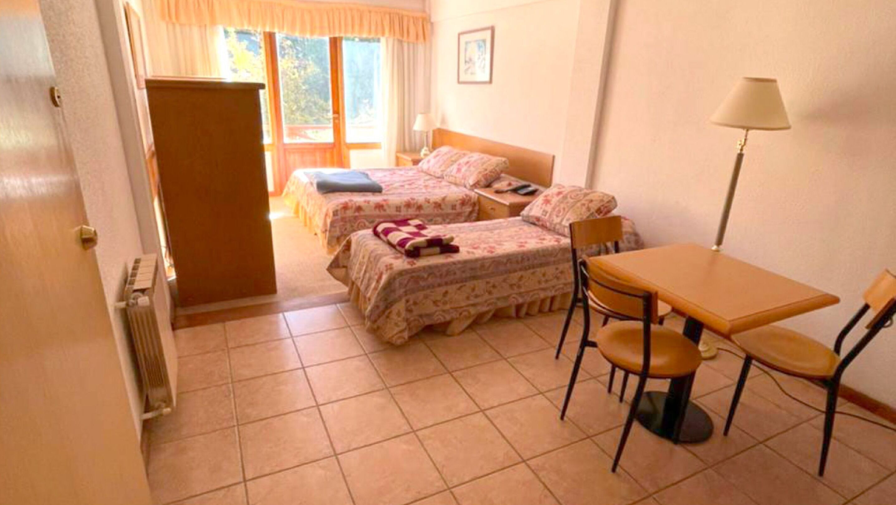 A room with two beds, a table, chairs, a lamp, and a radiator.