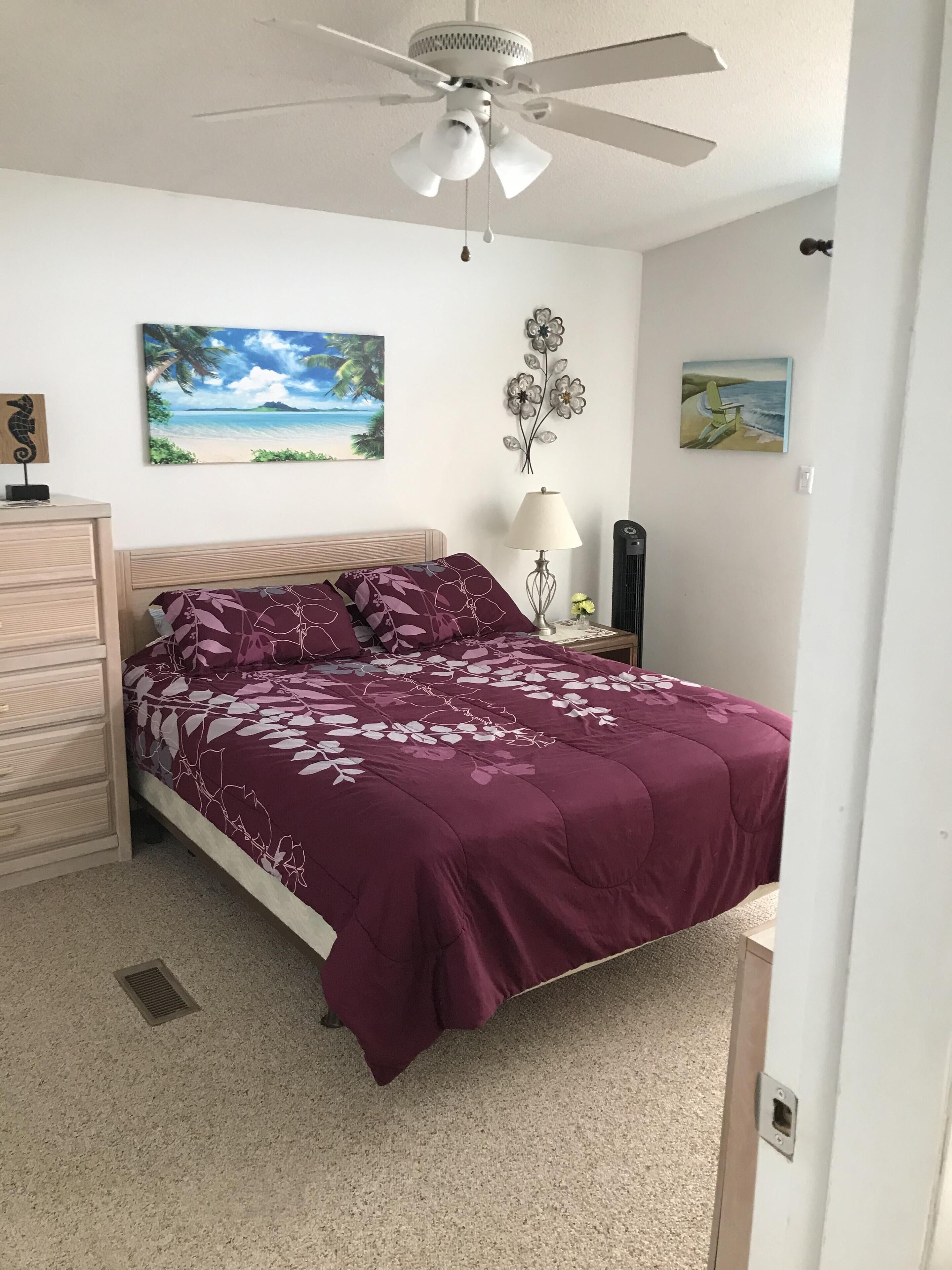 A bedroom with a bed, a ceiling fan, a nightstand, a lamp, and wall art.