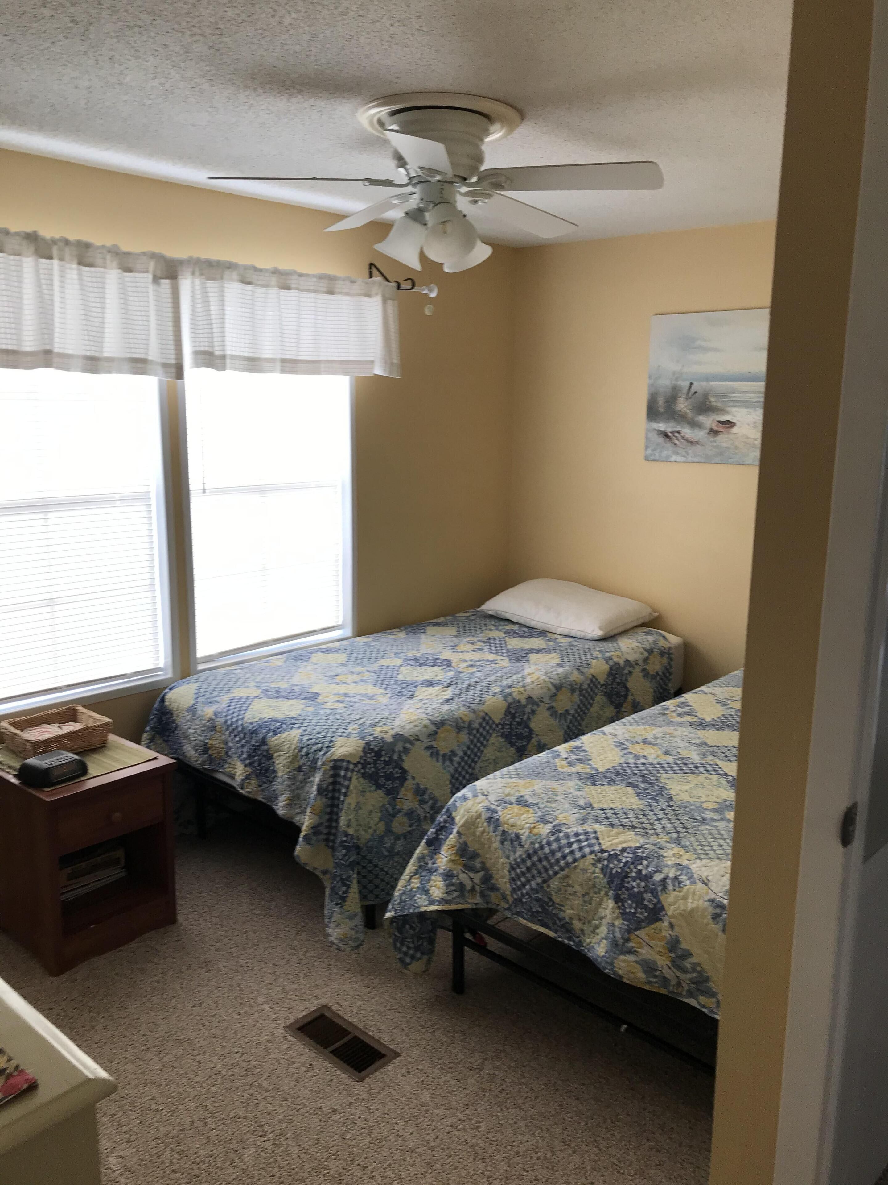 A small, well-lit room with two beds, a ceiling fan, a window with blinds, a painting on the wall, and a small desk with a basket.
