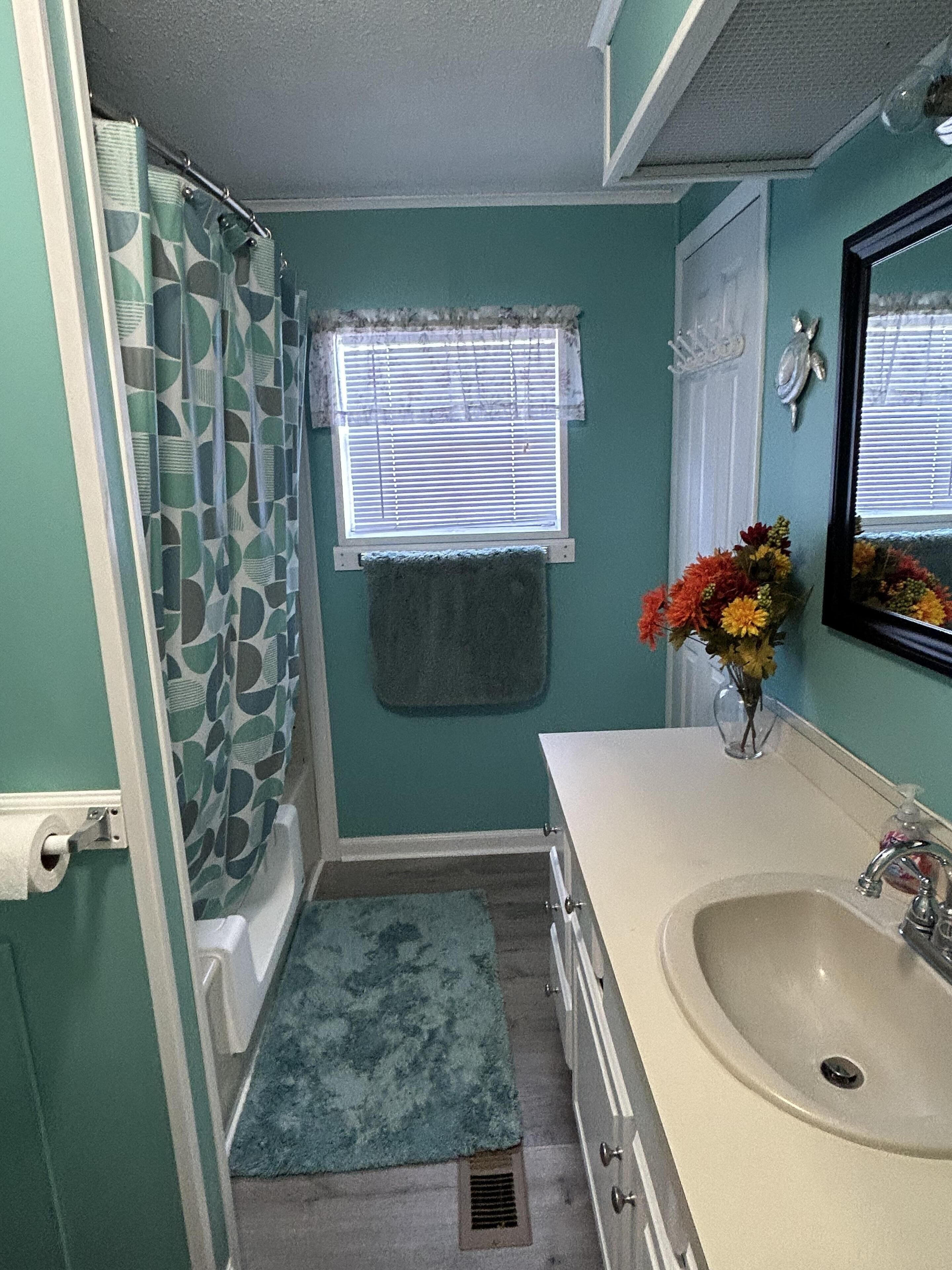 A bathroom with a bathtub, a sink, a window with blinds, a towel rack, and a vase with flowers.