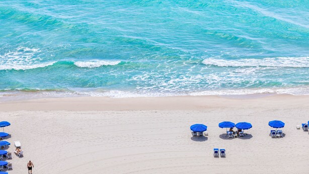 A beach with clear turquoise water, sandy shore, and several blue umbrellas and lounge chairs. - Private Oceanfront Condominium with Beach Access and Resort Services (Sunny Isles Beach)