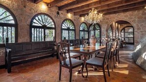 Un comedor rústico con una mesa y sillas de madera, paredes de piedra y grandes ventanales arqueados.