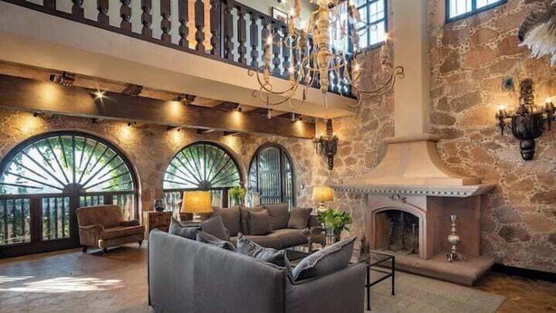Un amplio salón con un gran sofá modular, una chimenea y un balcón con paredes de piedra y ventanas arqueadas.