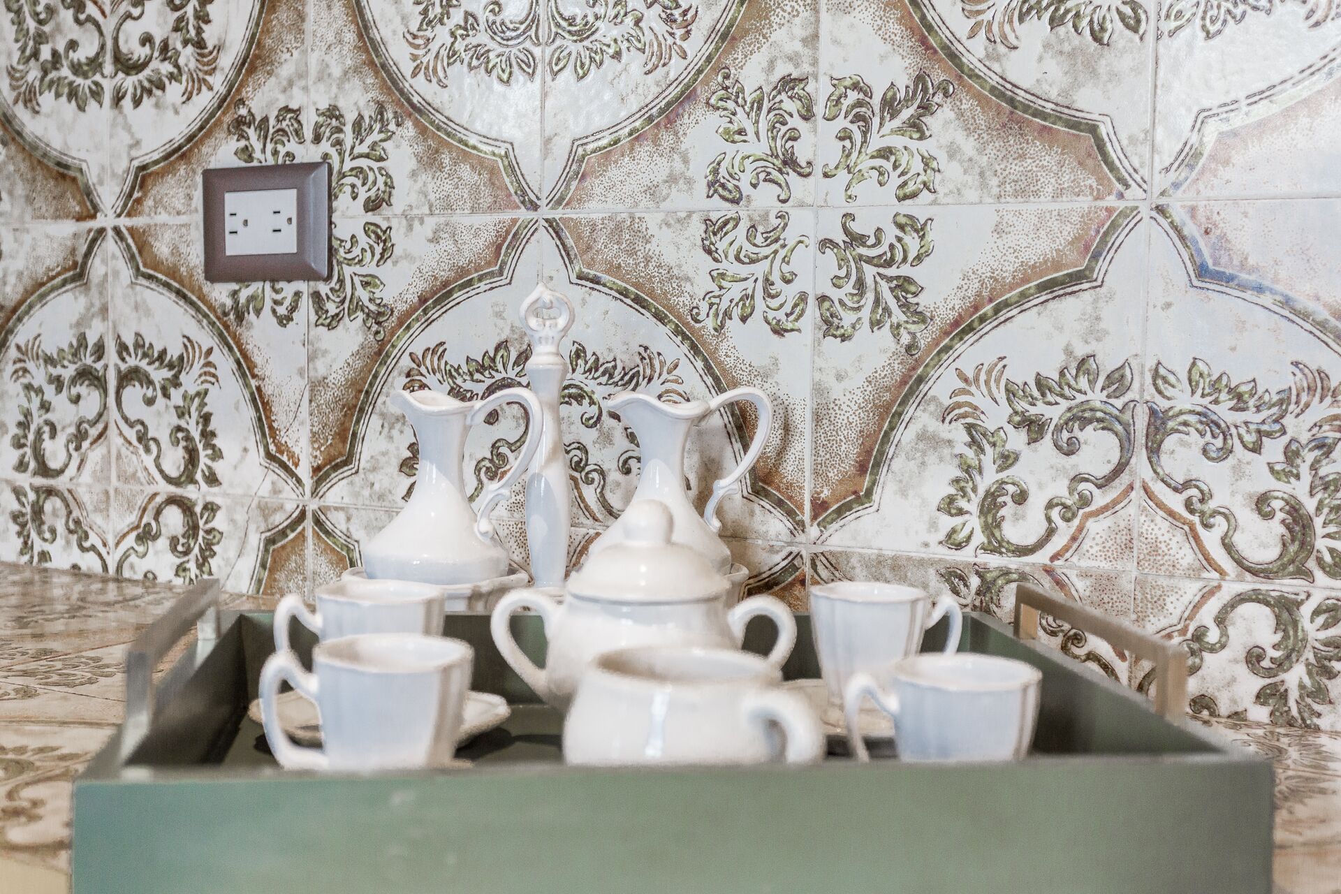 Una bandeja con teteras y tazas de cerámica blanca frente a una pared de azulejos decorados.