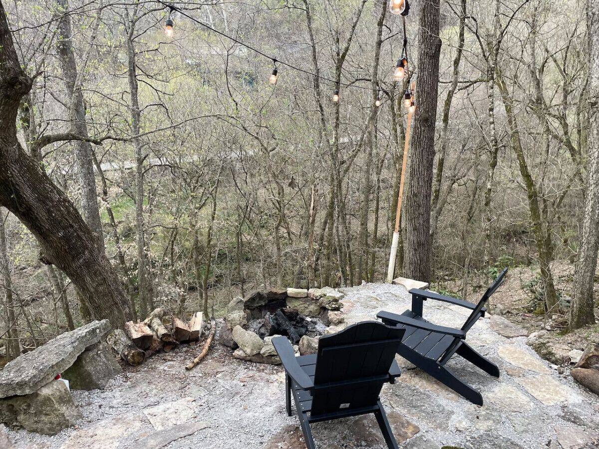 Un terrain de camping avec deux chaises noires, un foyer et des guirlandes lumineuses, le tout situé dans une zone boisée.
