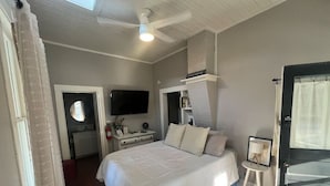 Un dormitorio con cama, televisor, ventilador de techo, una mesita y una puerta.