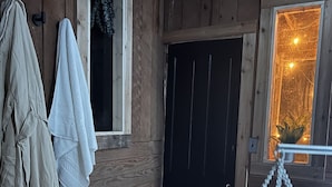 Interior de una cabaña de madera con una puerta negra, una toalla colgada y un cuenco metálico sobre un felpudo.