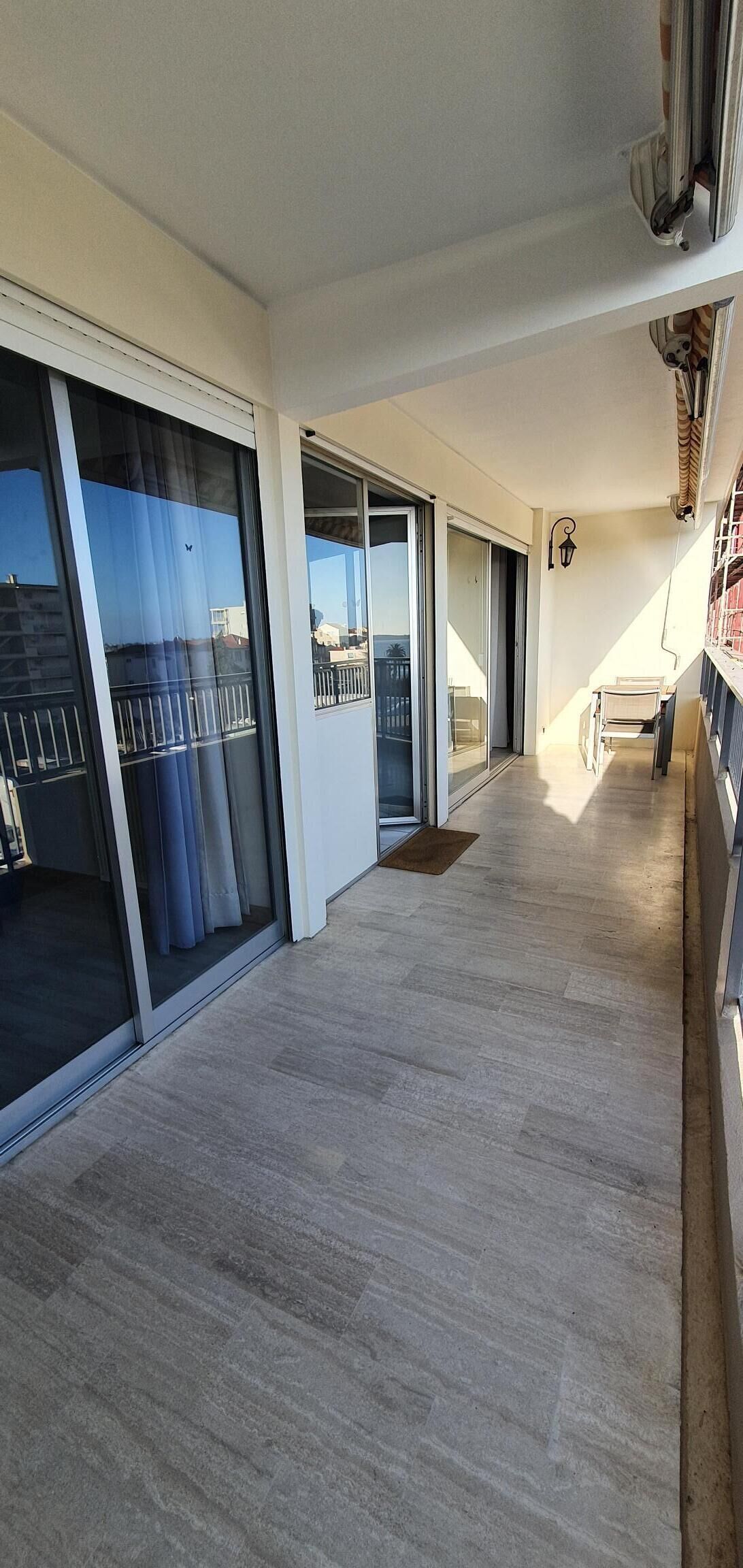 A corridor with a wooden floor, large sliding glass doors leading to a balcony, and a small seating area with a chair and table.