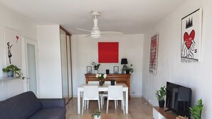 A modern living room with a grey sofa, a wooden coffee table, a dining area with a red accent wall, and a TV on a white cabinet. - Charming T2 48m² Balcony Sea View 4 sleeps 2min from the Beach (Vallauris)