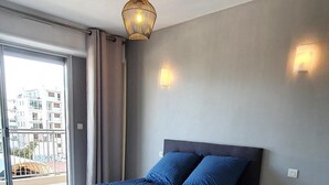 A bedroom with a large bed, a ceiling light, a wall-mounted lamp, and a view of the outside. - Charming T2 48m² Balcony Sea View 4 sleeps 2min from the Beach (Vallauris)