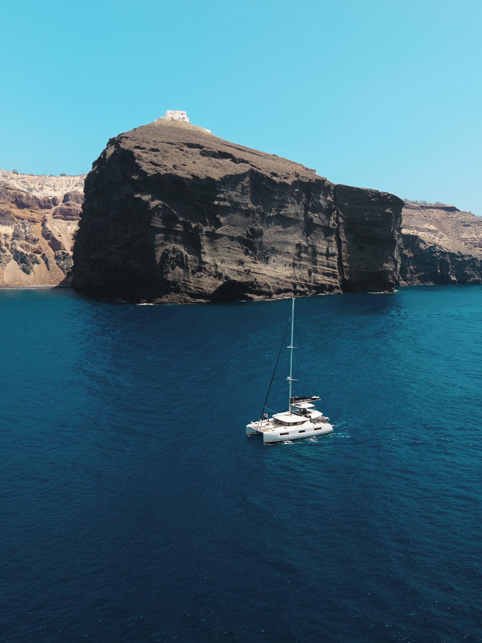 Un velero anclado cerca de una costa rocosa con un acantilado destacado y un pequeño edificio en la cima.