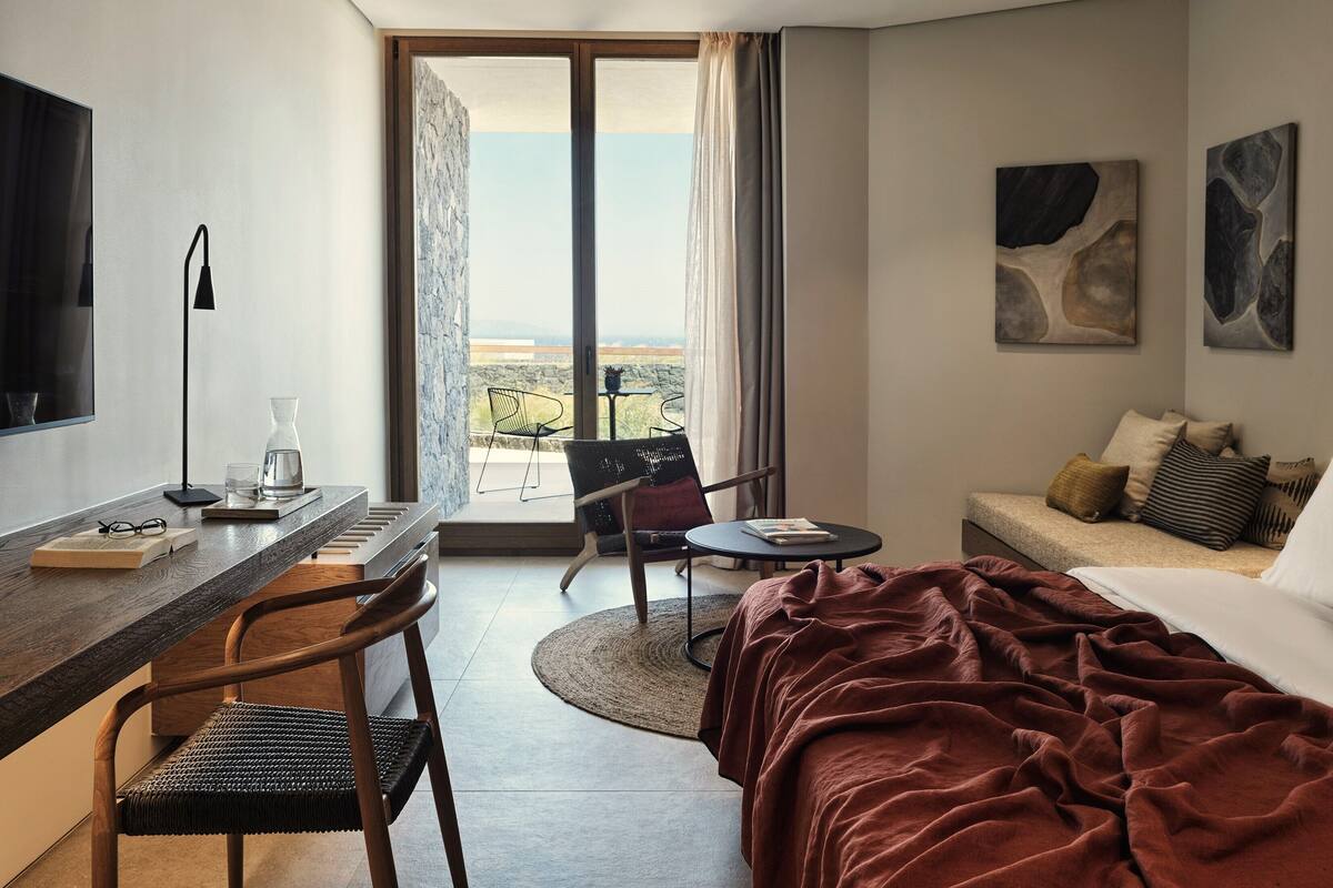 Ein modernes Hotelzimmer mit einem großen Bett, einem Schreibtisch mit Stuhl, einem kleinen Tisch mit Stuhl und einem Balkon mit Blick.