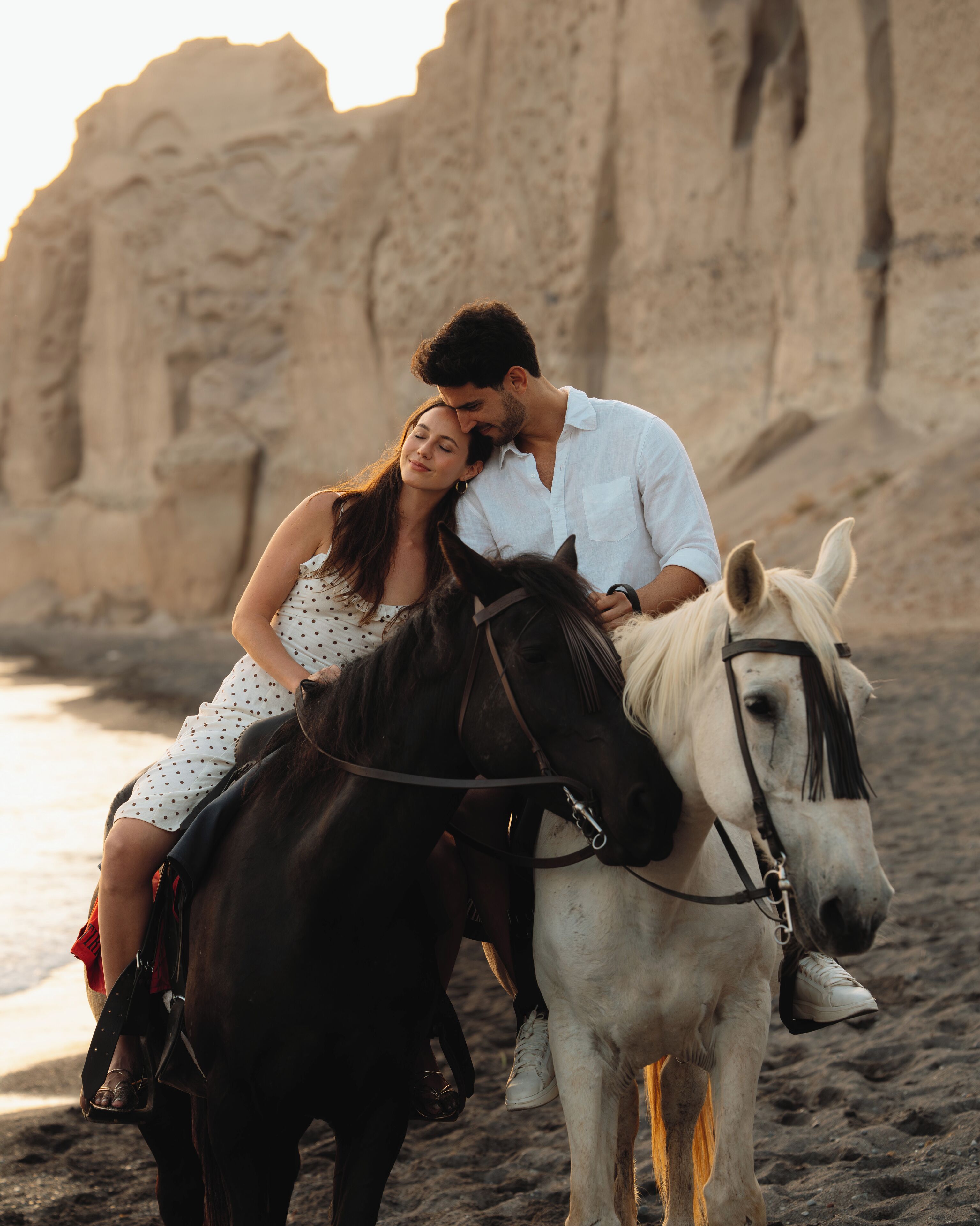 A couple riding horses on a beach with rocky cliffs in the background.