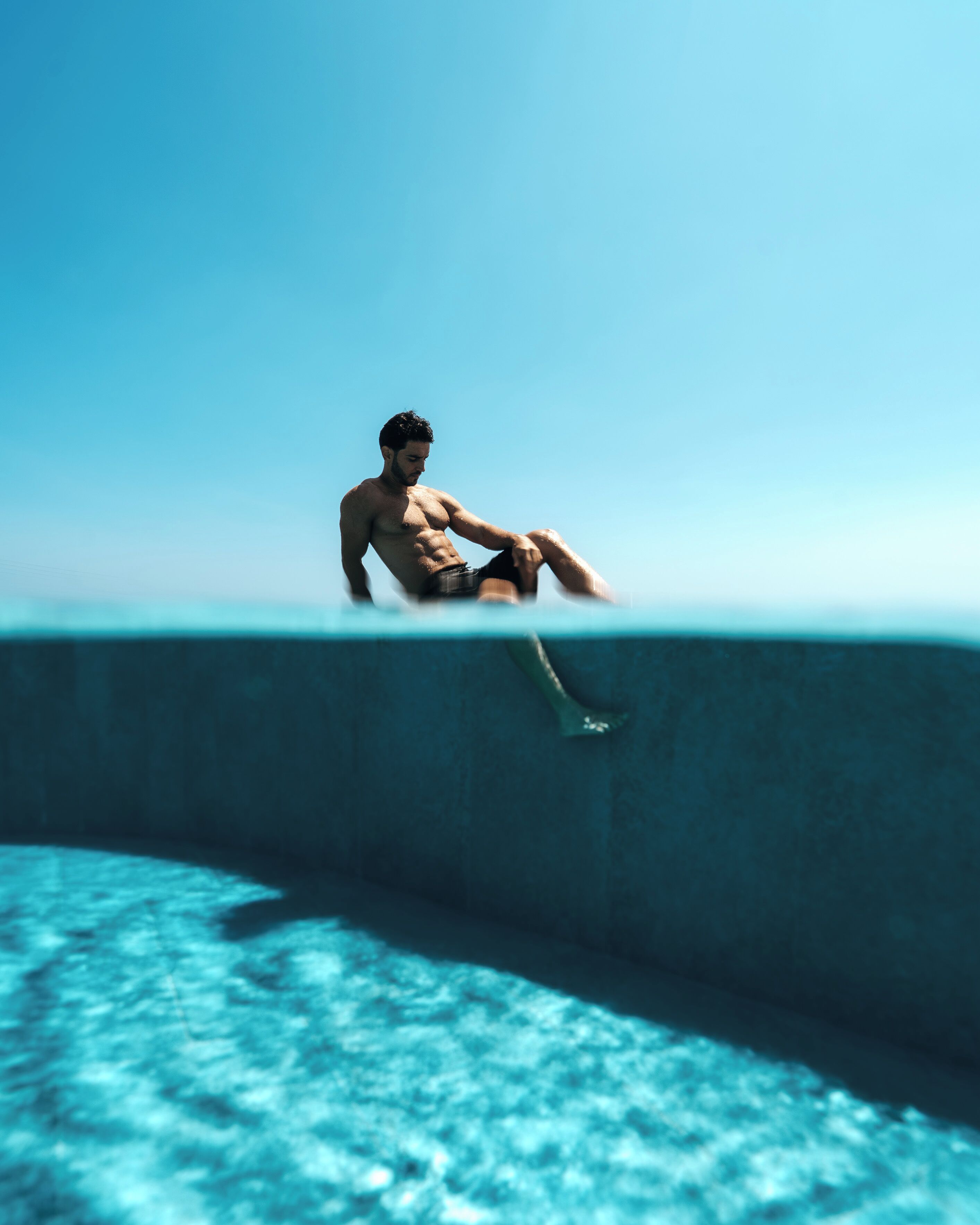A man sitting on the edge of a pool with a clear blue sky in the background.