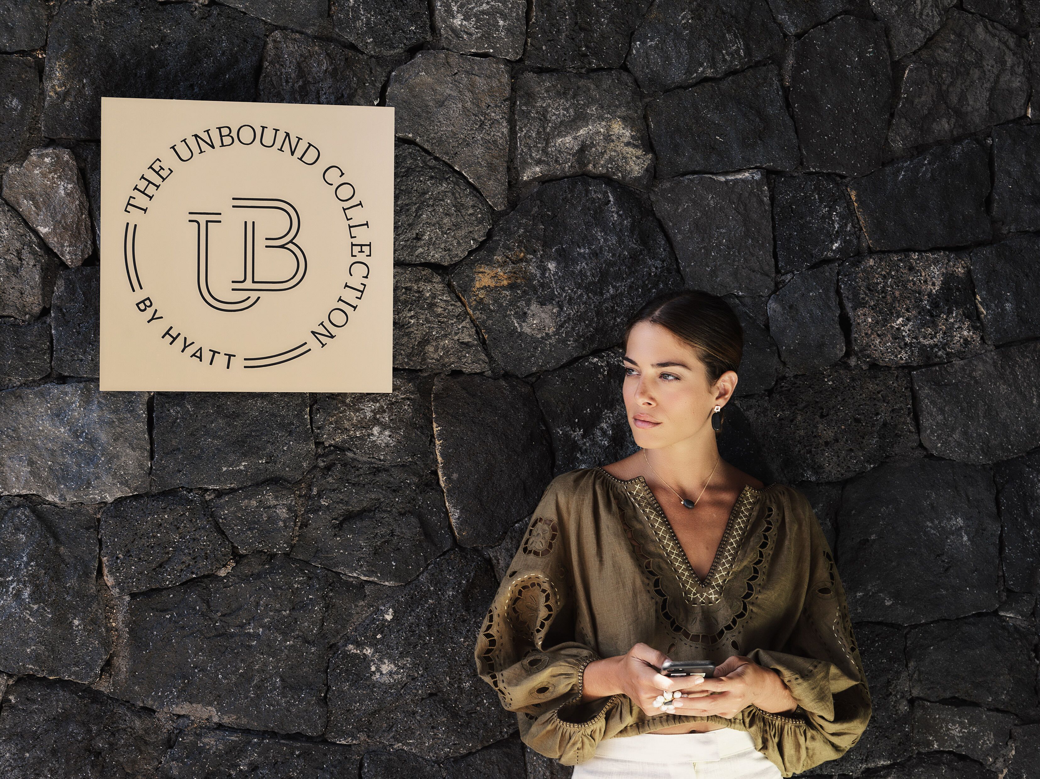 A woman standing in front of a stone wall with a sign that reads "THE UNBOUND COLLECTOR BY HYATT".