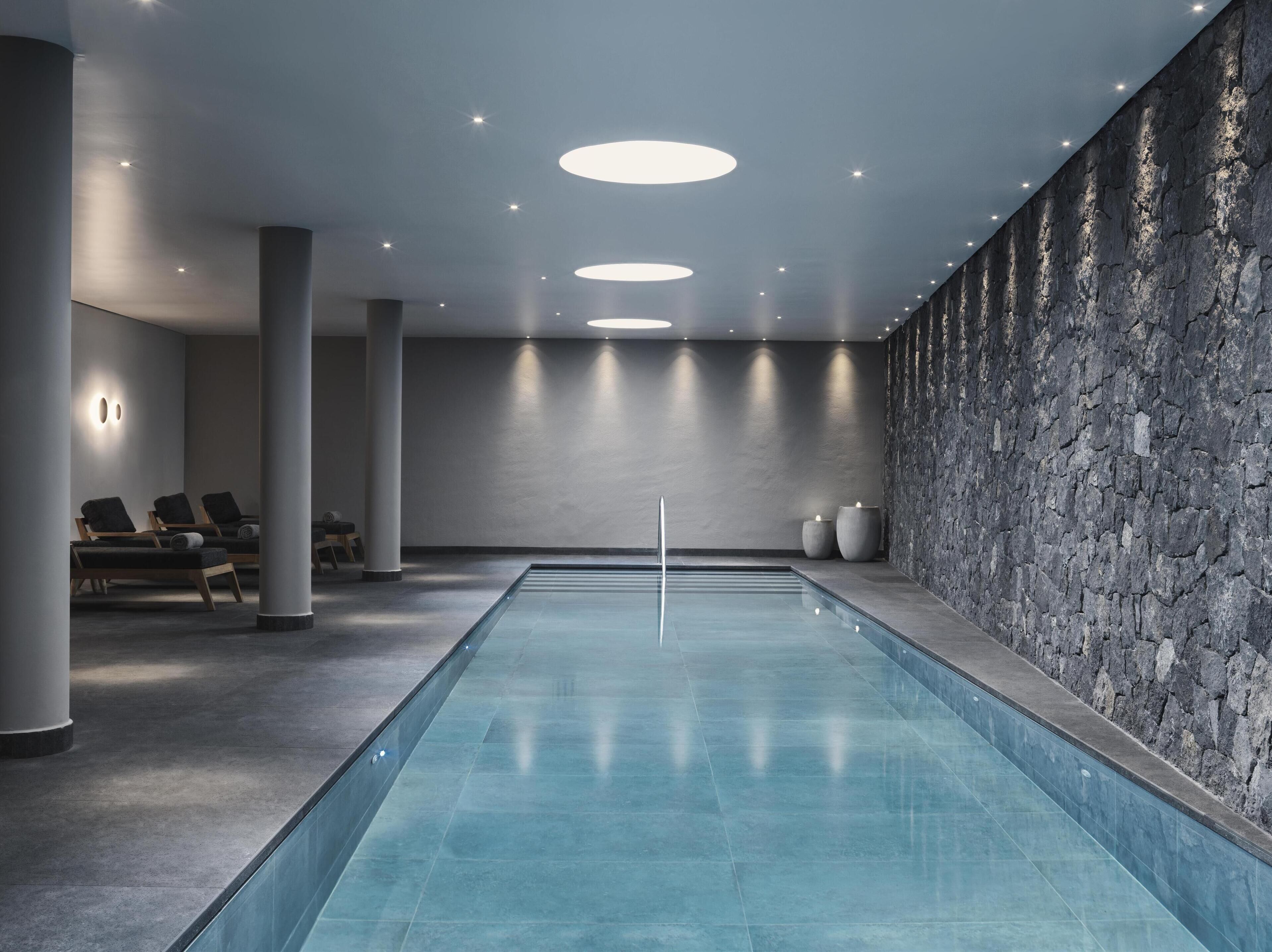 A modern indoor swimming pool with a stone wall and lounge chairs.