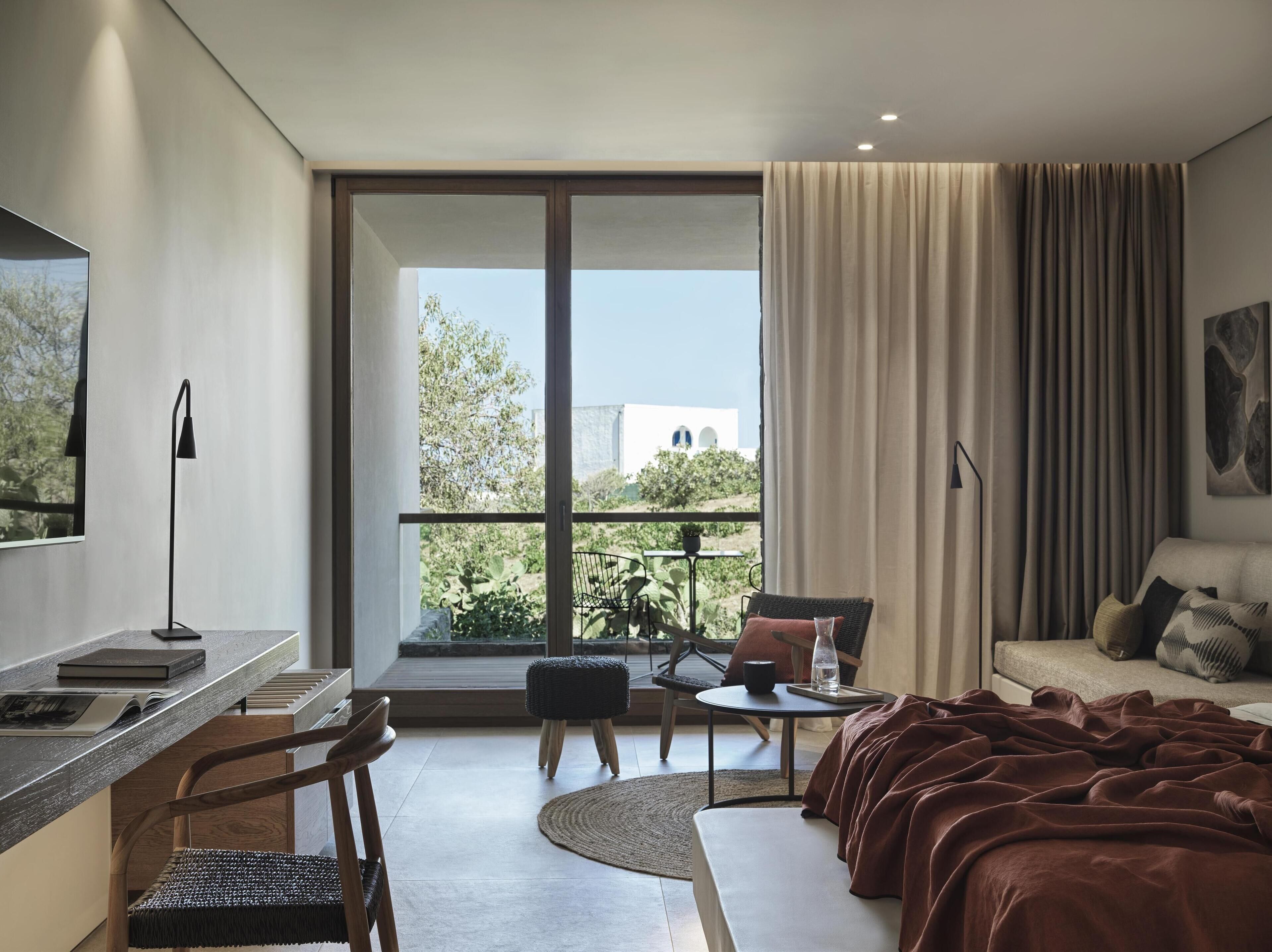 A modern hotel room with a large balcony, a sofa, a desk, and a chair.