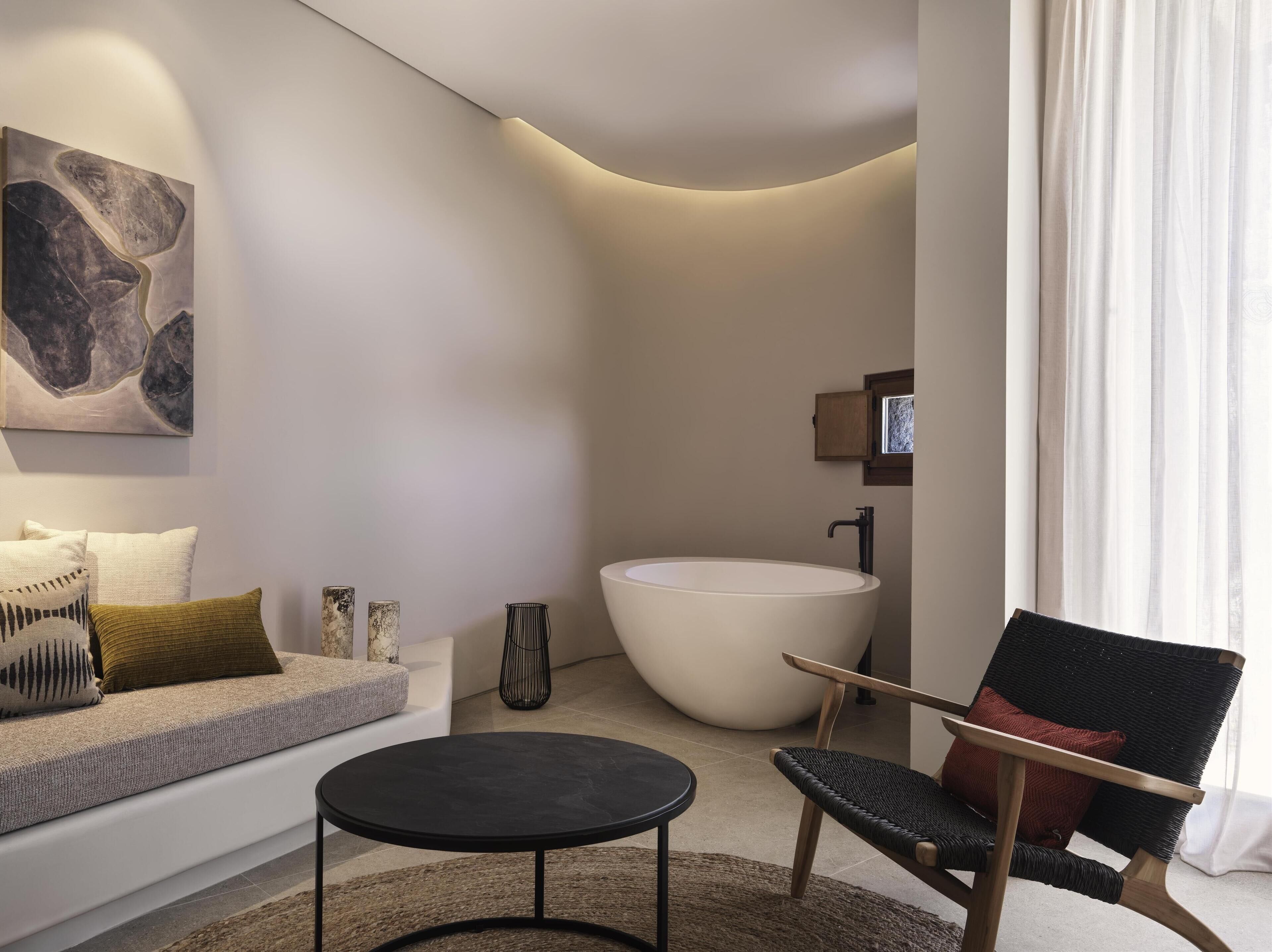 A modern living room with a sofa, a round black table, a wicker chair, a large white bathtub, and a minimalist artwork on the wall.