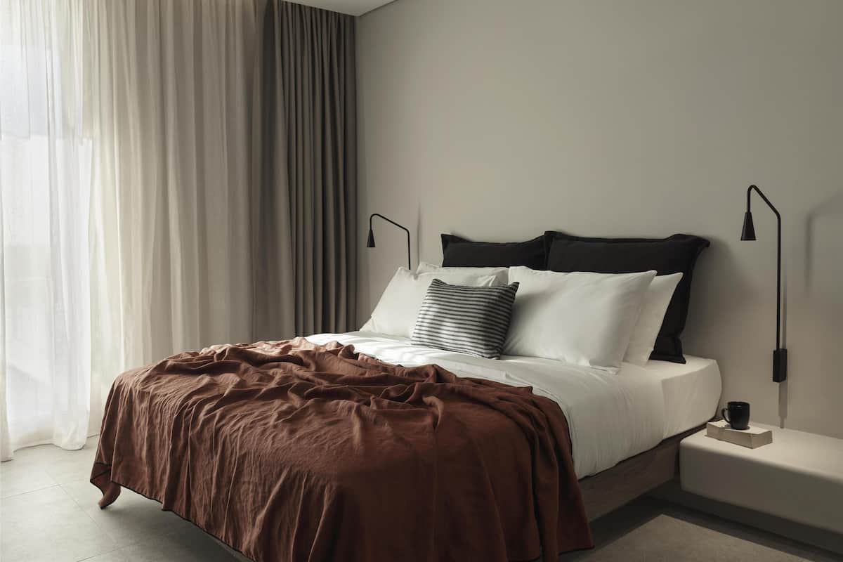 Ein modernes Schlafzimmer mit einem großen Bett, zwei Nachttischen, einem kleinen Tischchen und einem Fenster mit transparenten Vorhängen.