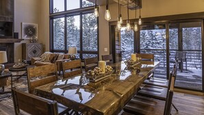 Un comedor amplio con una gran mesa de madera y numerosas sillas, una chimenea y ventanas grandes que ofrecen vistas a árboles cubiertos de nieve.