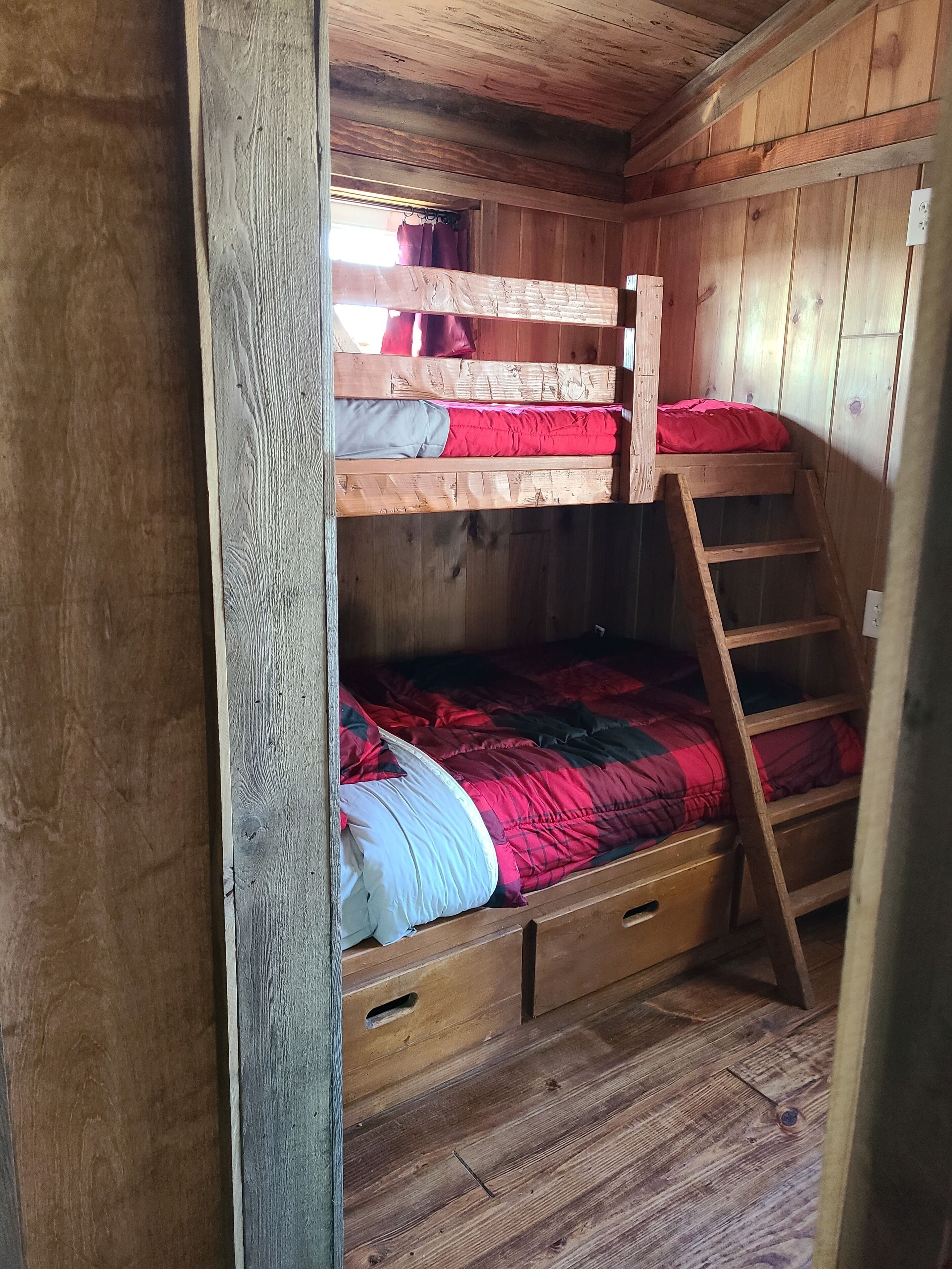 A bunk bed with a ladder, wooden walls, and a window with curtains.