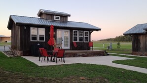A rustic wooden building with a metal roof, a covered patio with red chairs, and a view of a green field. - Cute Family Cabin at Lakeview by Sue  (Appleton City)