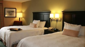 Quarto de hotel com duas camas, uma mesa de cabeceira com abajur, uma cadeira e um quadro na parede.