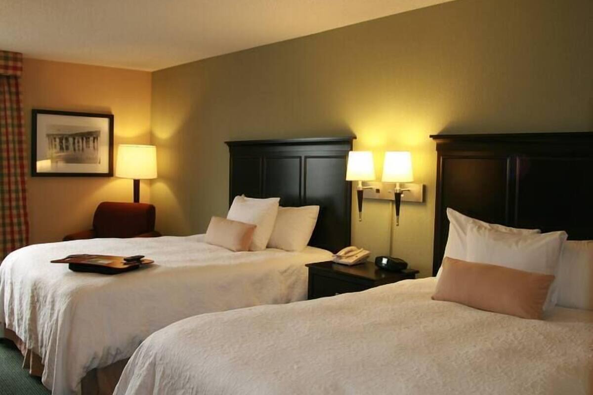 Habitación de hotel con dos camas, una mesita de noche con lámpara, una silla e imagen enmarcada en la pared.