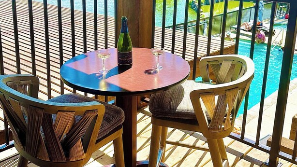 Um deck de madeira com uma mesa redonda e duas cadeiras de vime, com vista para uma piscina e um rio.