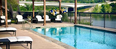 Zona de piscina con sillones de descanso y una pérgola, con vista a un puente y vegetación.
