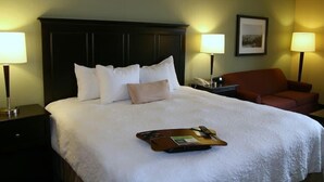 Quarto de hotel com uma cama grande, duas mesas de cabeceira, uma cadeira e um suporte com um controle remoto e um livro.