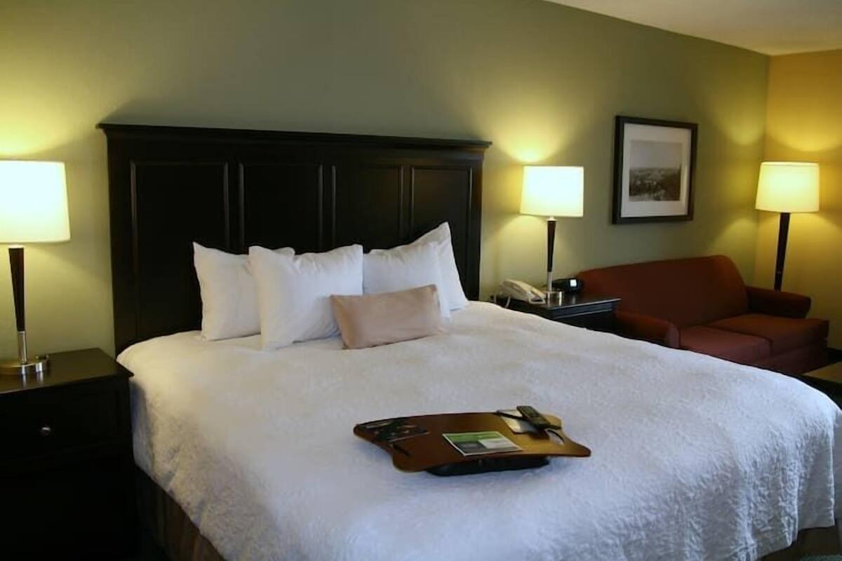 Habitación de hotel con una cama grande, dos mesitas de noche, una silla y una bandeja con un control remoto y un libro.
