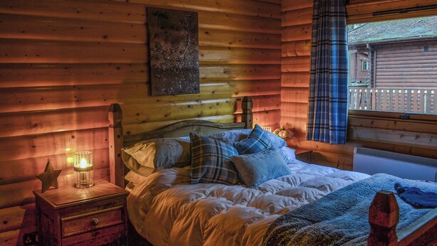 A cozy wooden cabin bedroom with a bed, a nightstand, a lamp, and a window with curtains. - Kenwick Lodge - UK37416 (Kenwick, near Louth)