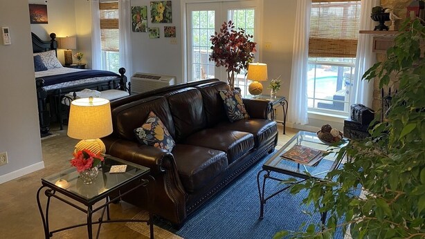 A living room with a leather sofa, a glass coffee table, and a lamp. There is a bed in the background and a window with curtains. - Cozy Cottage with Private pool, Private Hot Tub and Internet (Wimberley)