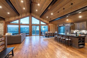 A spacious kitchen with a large island, wooden flooring, and large windows offering a view of the outdoors.
