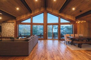 A spacious room with a wooden ceiling, large windows, a stone wall, a sofa, and a dining table. - Ponderosa Peak. Games. Views. BBQ. Hammock. (Big Bear Lake)
