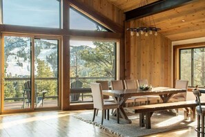 A wooden interior with a large dining table, chairs, and a view of a snowy landscape through large windows.