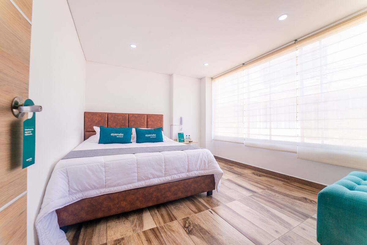 A modern hotel room with a wooden floor, a large bed with white bedding and teal pillows, a wooden headboard, a teal armchair, and a window with blinds.