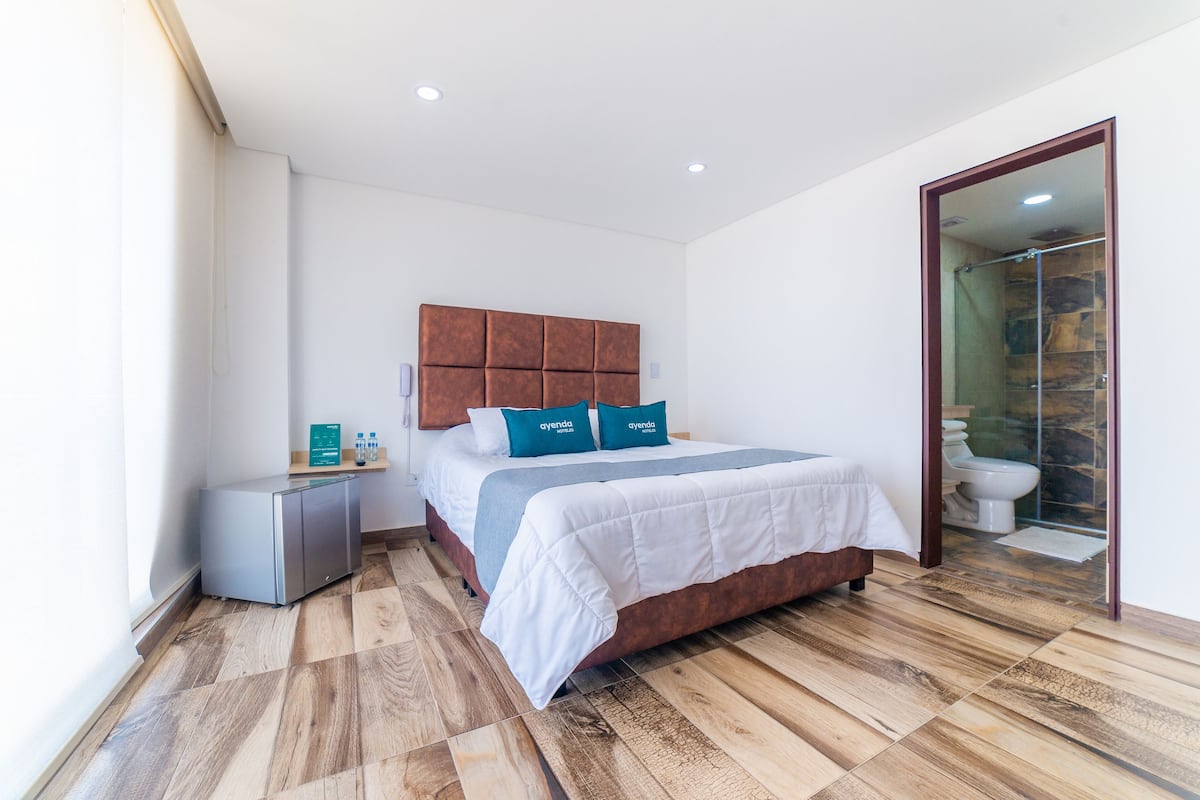 A modern bedroom with a wooden floor, a bed with white bedding and teal pillows, a small bedside table, and a glass shower enclosure.
